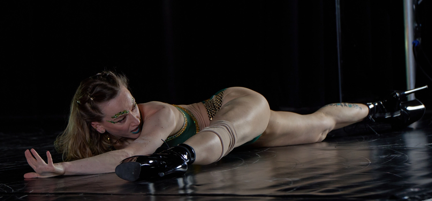 Polina, professeure de pole dance exotique au Momentum Studio Strasbourg, réalise une figure au sol mêlant puissance et sensualité sur une scène sombre éclairée.