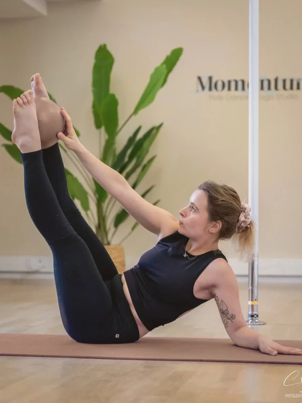 Élève en exercice de pilates sur tapis, jambes tendues et ballon entre les pieds, dans le studio Momentum à Strasbourg avec barre de pole dance en fond