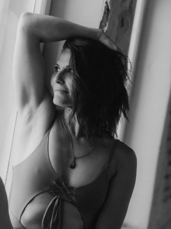 Portrait en noir et blanc de Soledad, professeure à Momentum Studio, bras levé contre une fenêtre, en body lacé et collier à pendentif
